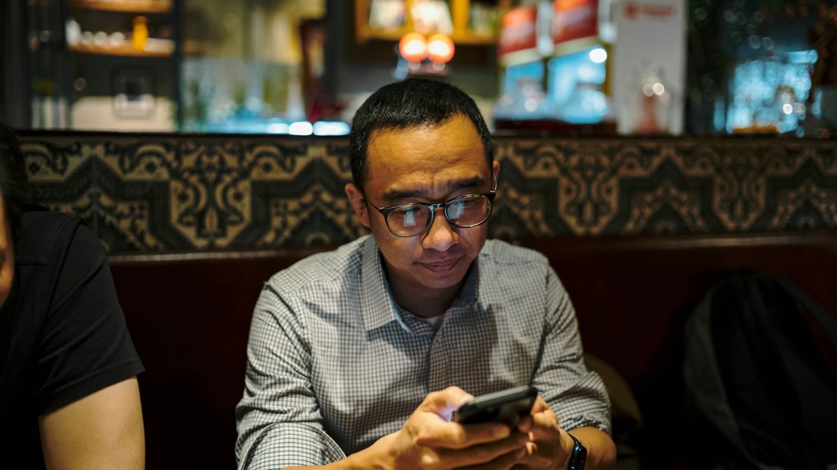 A man in glasses reading his phone at a restaurant table, the bar lit behind him
