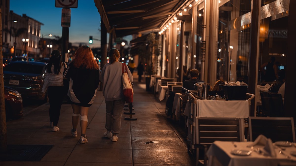 People walking past lit restaurant windows on a city street at dusk