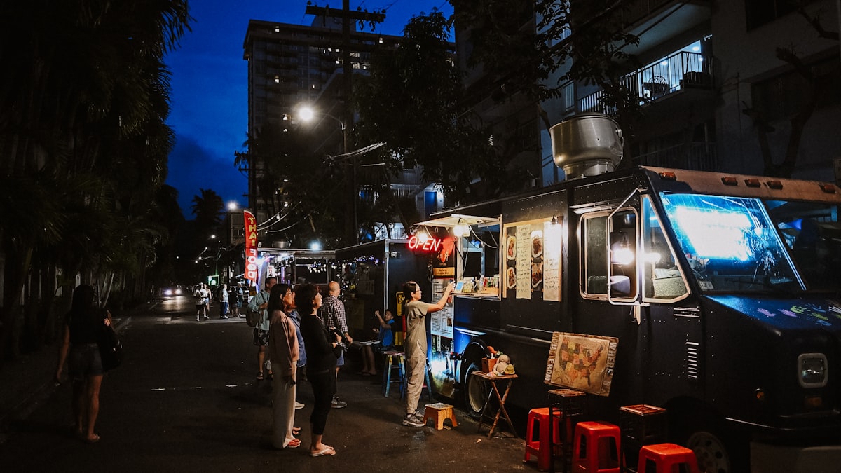 Customers ordering at a lit food truck on a dark city street, red stools on the pavement